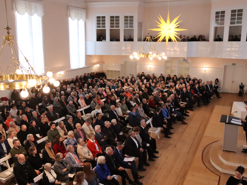 Ein großer heller Saal ist gefüllt mit Menschen, die auf Bänken sitzen und einem Redner vorne zuhören.