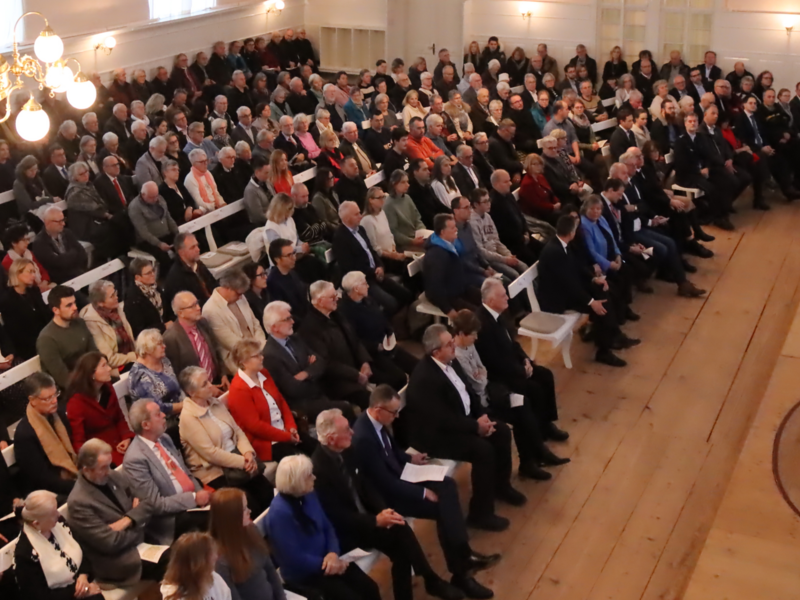 Ein großer heller Saal ist gefüllt mit Menschen, die auf Bänken sitzen und einem Redner vorne zuhören.