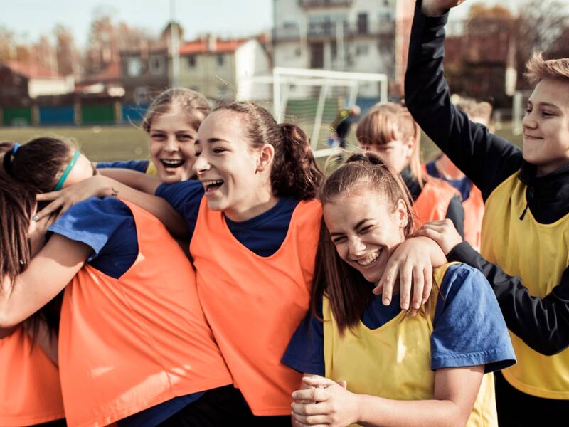 Eine Gruppe von Jugendlichen in Sportkleidung lacht und umarmt sich auf einem Fußballfeld.