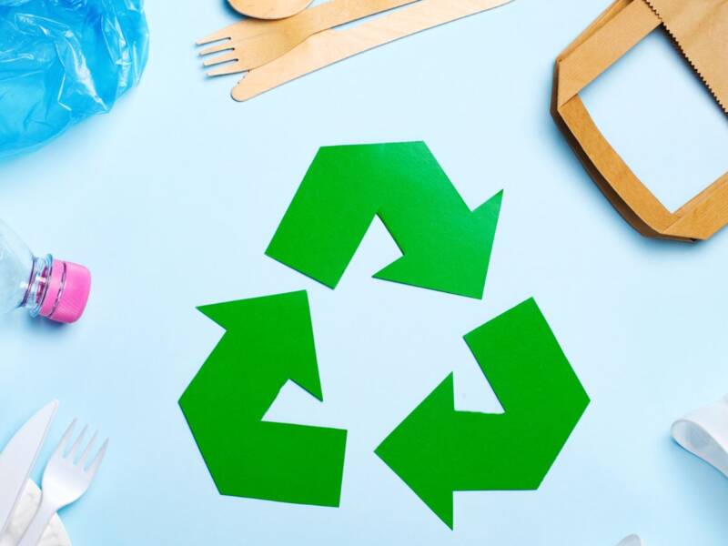 Auf einem hellblauen Hintergrund liegt das grüne Recycling-Symbol, umgeben von verschiedenen Einwegverpackungen aus Plastik, Papier und Holz.