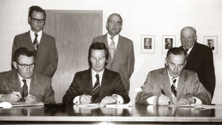 Schwarz-weiß Bild von 1975 mit 6 Männern im Anzug, drei davon sitzen an einem langen Schreibtisch, die anderen drei stehen dahinter. Die sitzenden Männern haben alle einen Stift in der Hand und Papier vor sich liegen.