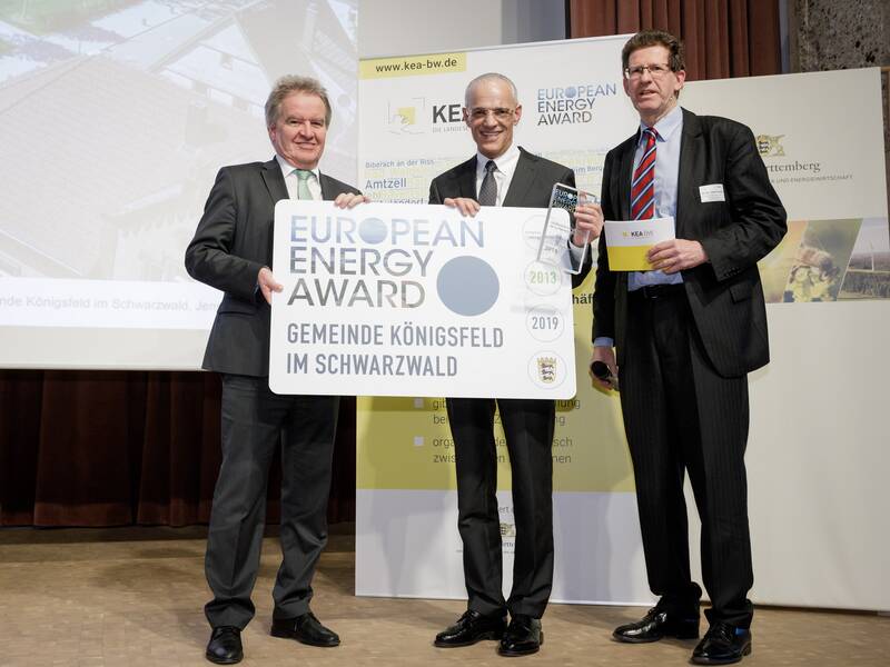 Drei Männer in Anzügen präsentieren gemeinsam ein Schild mit der Aufschrift "European Energy Award - Gemeinde Königsfeld im Schwarzwald" vor einem Plakat und einer Leinwand.