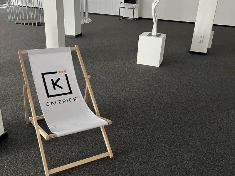 Ein heller Liegestuhl mit der Aufschrift "GALERIE K3" befindet sich im Vordergrund eines modern eingerichteten Kunstgalerieraums mit Skulpturen und Bildern an den Wänden.