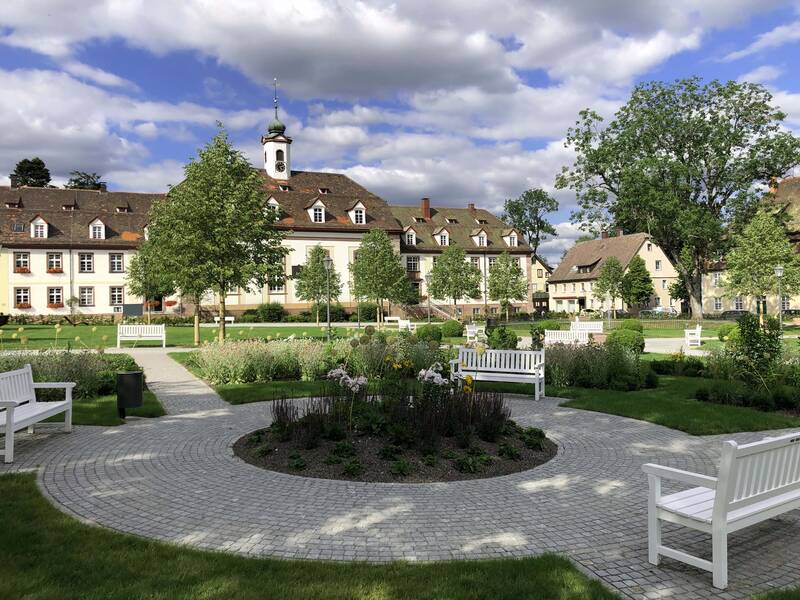 Ein gepflegter Park mit weißen Bänken und Blumenbeeten liegt vor einem historischen Gebäude unter blauem Himmel mit Wolken.