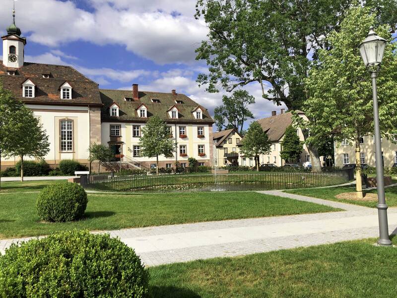 Ein gepflegter Park mit einem Springbrunnen und historischen Gebäuden im Hintergrund an einem sonnigen Tag.