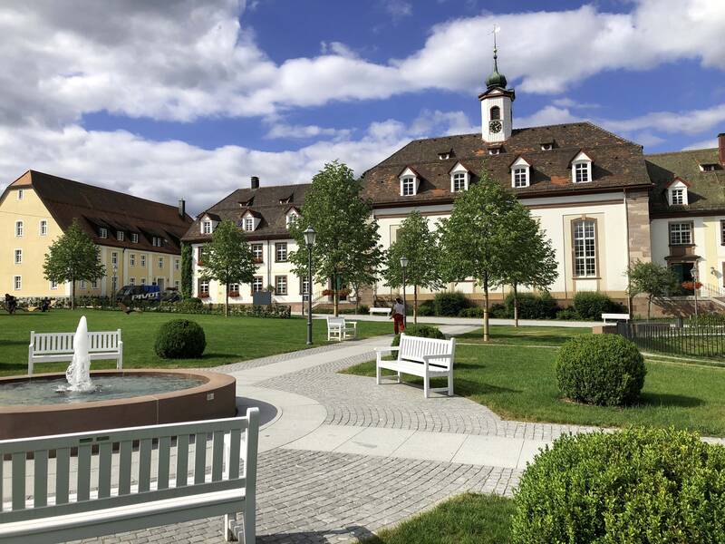 Ein malerischer Park mit weißen Bänken, einem runden Brunnen und einem historischen Gebäude im Hintergrund unter blauem Himmel mit Wolken.