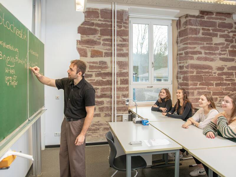 Ein Lehrer steht vor einer grünen Tafel und erklärt einer Gruppe von vier Schülerinnen eine mathematische Gleichung.