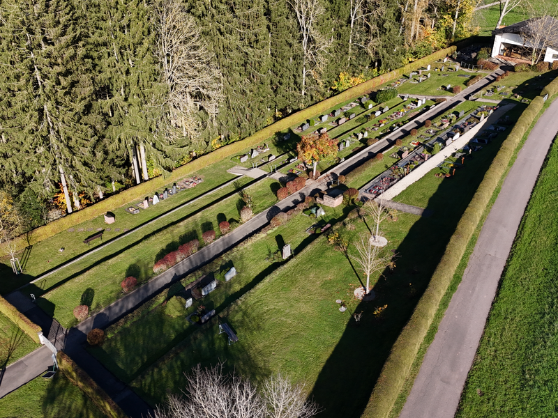 Eine Luftaufnahme zeigt einen gepflegten Friedhof mit mehreren Grabreihen, umgeben von Bäumen und einem angrenzenden Weg.