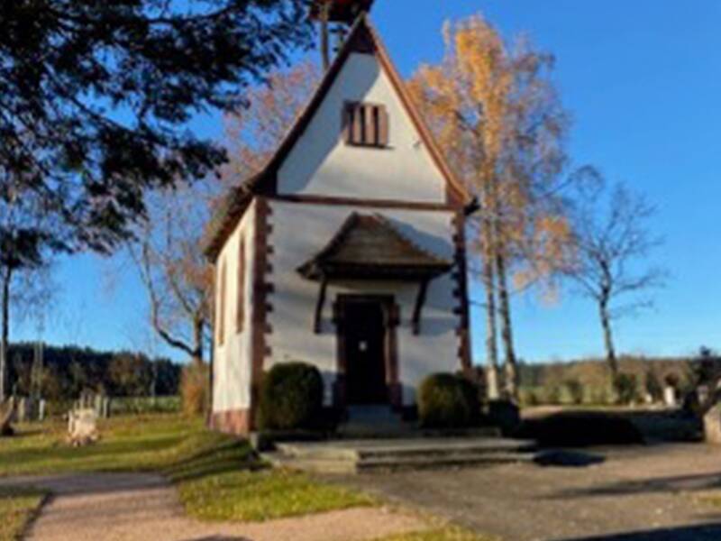 Eine kleine Kapelle auf einer Anhöhe vor blauem Himmel mit kahlen Bäumen im Vordergrund