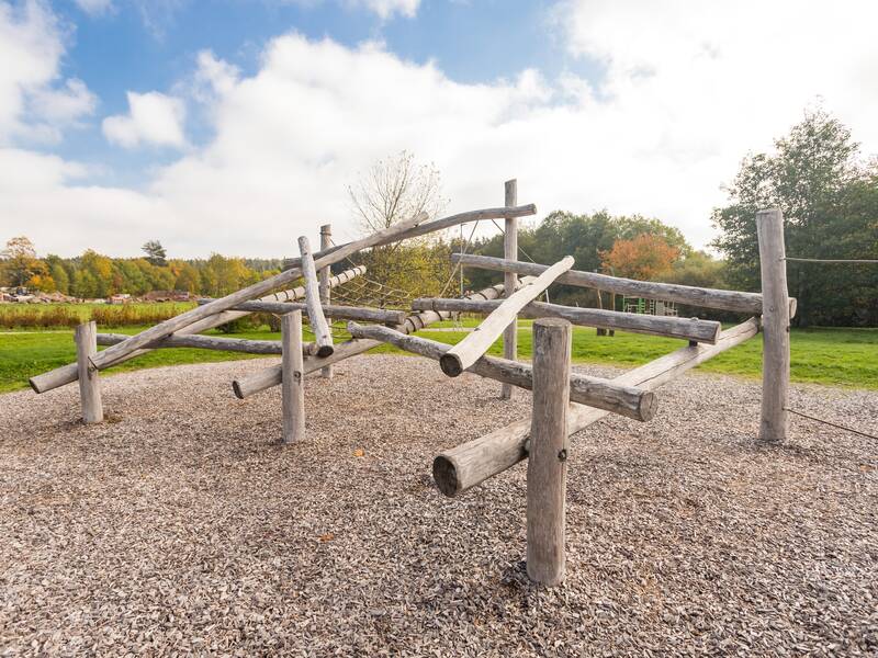 Ein Klettergerüst aus Holzstämmen steht auf einem Kiesbett in einem weitläufigen, grünen Park unter bewölktem Himmel.