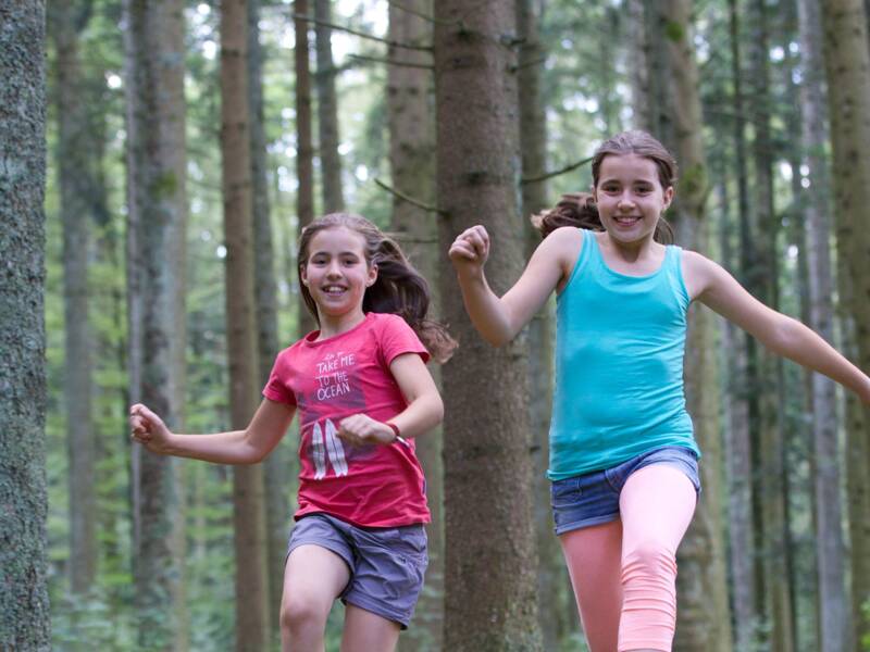 Zwei Kinder rennen gemeinsam durch einen Wald und springen dabei in die Luft.