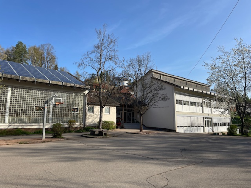 Ein Schulgebäude unter klarem Himmel, ausgestattet mit Solarpaneelen auf einem Dachteil und einem Basketballkorb vorne.