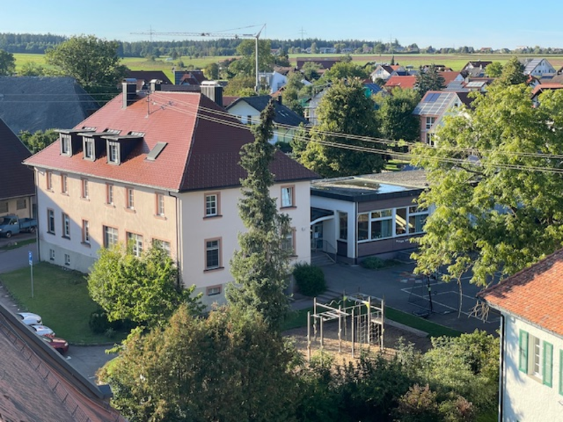 Ein Blick von oben auf ein Schulgebäude mit einem roten Dach, das an einen Spielplatz und eine Straße angrenzt, mit einer ländlichen Landschaft im Hintergrund.