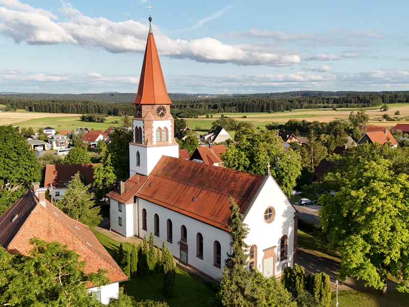 Eine Kirche mit rotem Ziegeldach und Turm steht inmitten einer idyllischen ländlichen Landschaft mit vielen Bäumen und vereinzelten Häusern im Hintergrund.