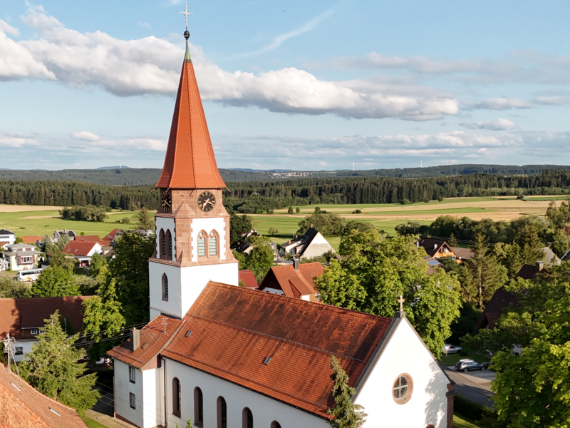 Eine Kirche mit rotem Ziegeldach und Turm steht inmitten einer idyllischen ländlichen Landschaft mit vielen Bäumen und vereinzelten Häusern im Hintergrund.