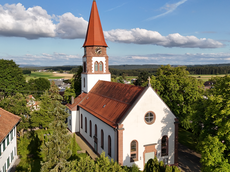 Eine Kirche mit einem roten Ziegeldach und einem Uhrenturm steht umgeben von Bäumen und einem weiten Landschaftsausblick unter einem blauen Himmel mit Wolken.