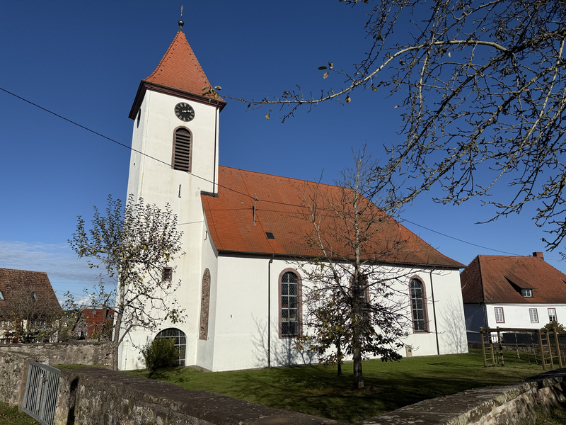 Eine weiße Kirche mit einem roten Ziegeldach und einem Turm steht auf einer grünen Wiese vor einem klaren blauen Himmel.