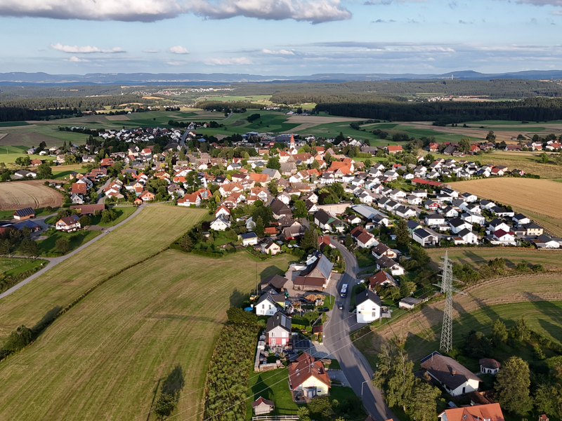 Die Luftaufnahme zeigt ein Dorf, umgeben von weiten Feldern und Waldregionen unter blauem Himmel.