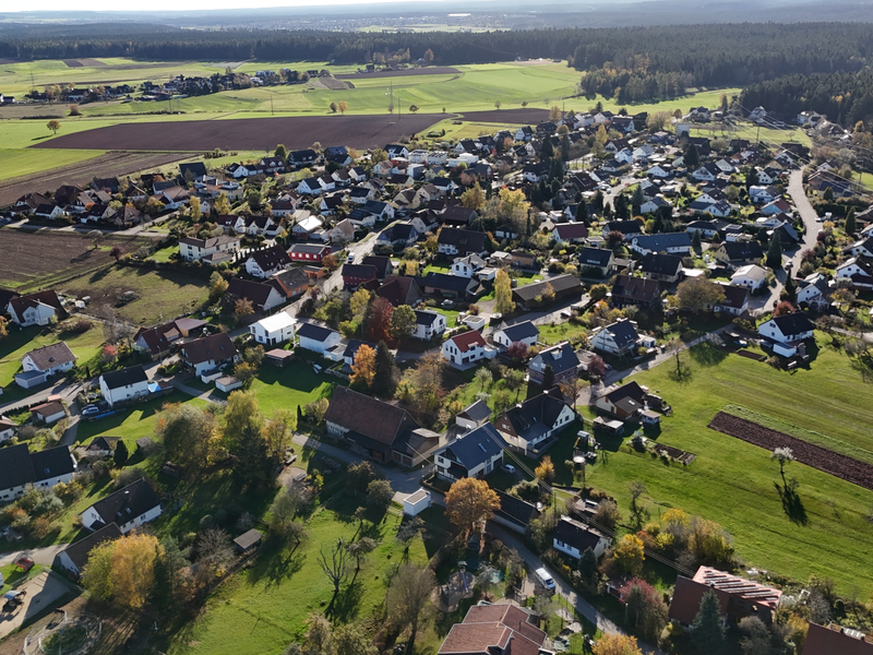 Die Luftaufnahme zeigt ein Dorf mit zahlreichen Häusern, umgeben von Feldern und Waldgebieten.