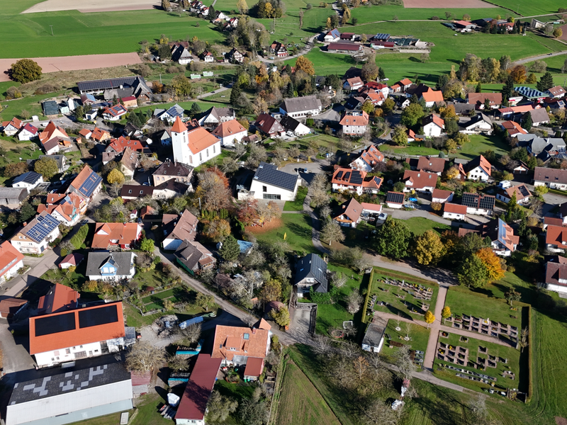 Die Luftaufnahme zeigt ein kleines Dorf mit vielen rotgedeckten Häusern, einer Kirche und einem angrenzenden Friedhof, umgeben von grünen Feldern und Wiesen.