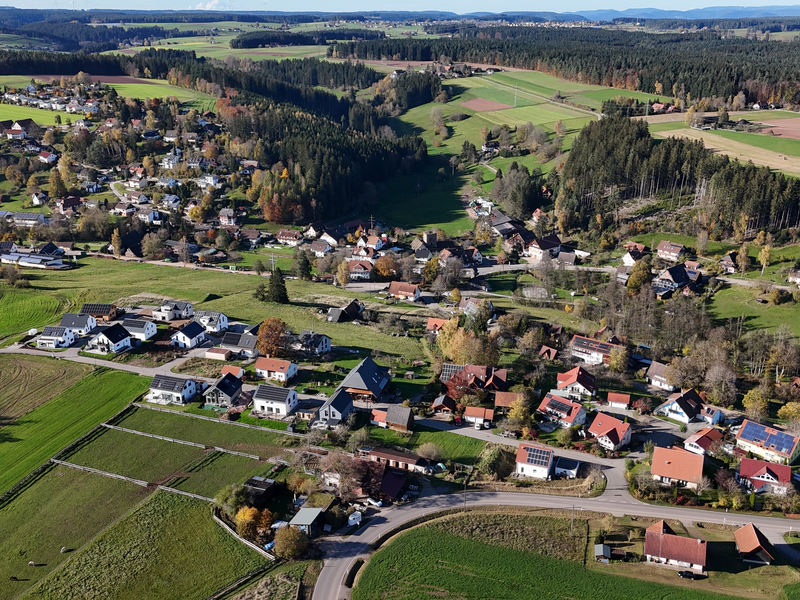 Eine Luftaufnahme zeigt eine kleine ländliche Siedlung umgeben von grünen Feldern und Waldgebieten.