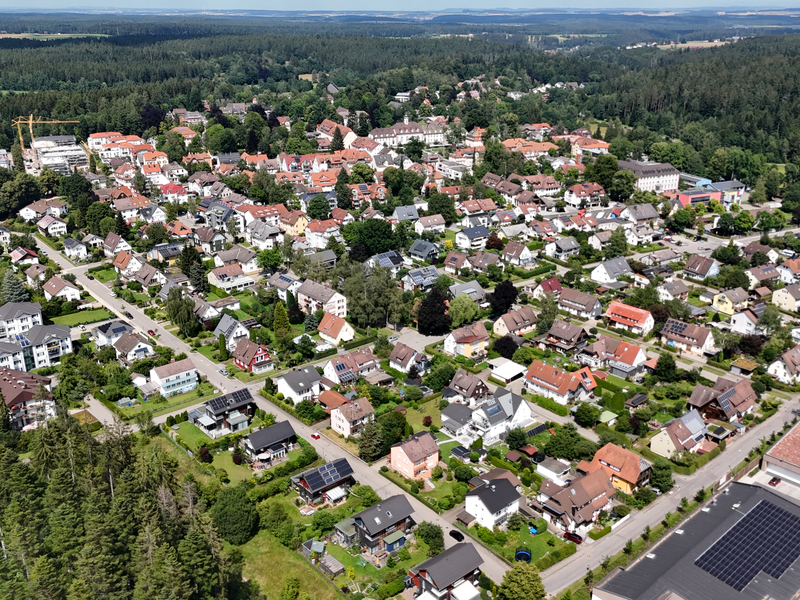 Luftaufnahme einer grünen Wohngegend mit vielen dicht beieinander stehenden Häusern, umgeben von Waldflächen.