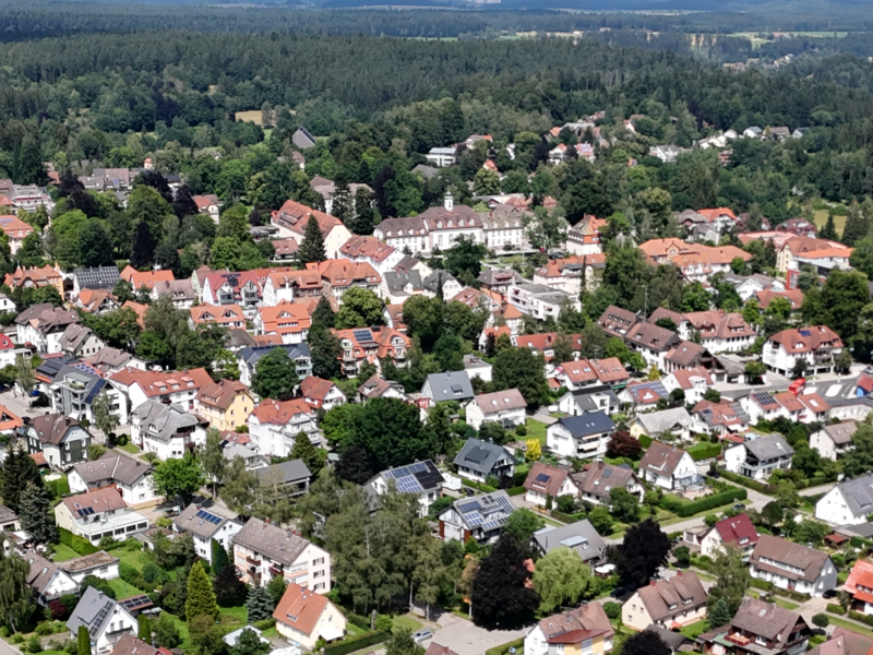 Luftaufnahme einer grünen Wohngegend mit vielen dicht beieinander stehenden Häusern, umgeben von Waldflächen.