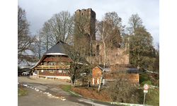 Ein traditionelles, rustikales Gebäude steht vor den Überresten einer alten Burg in einer bewaldeten Landschaft.