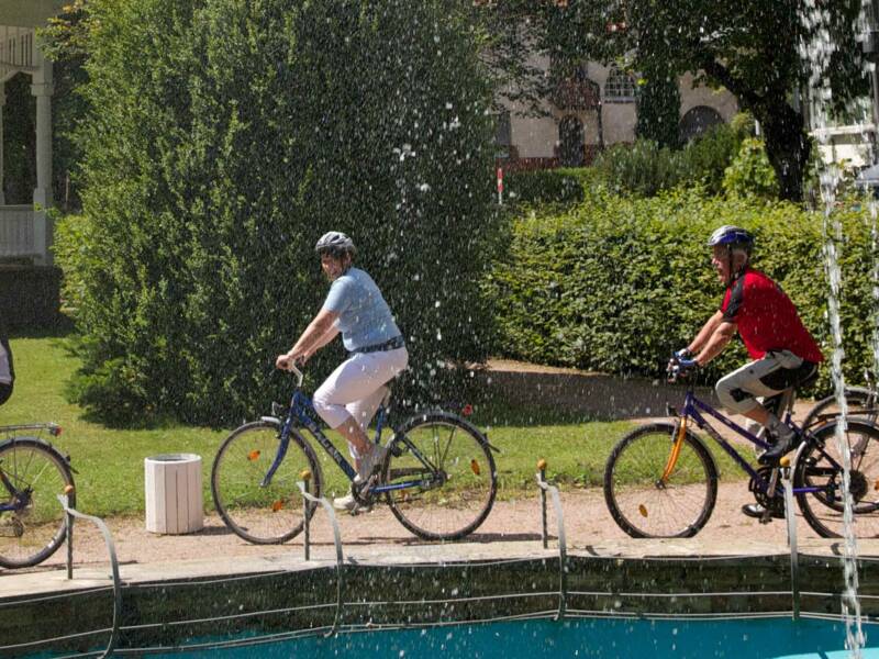 Eine Gruppe von Personen fährt auf Fahrrädern auf einem Weg an einem Springbrunnen vorbei, umgeben von Bäumen und einem großen Gebäude im Hintergrund.