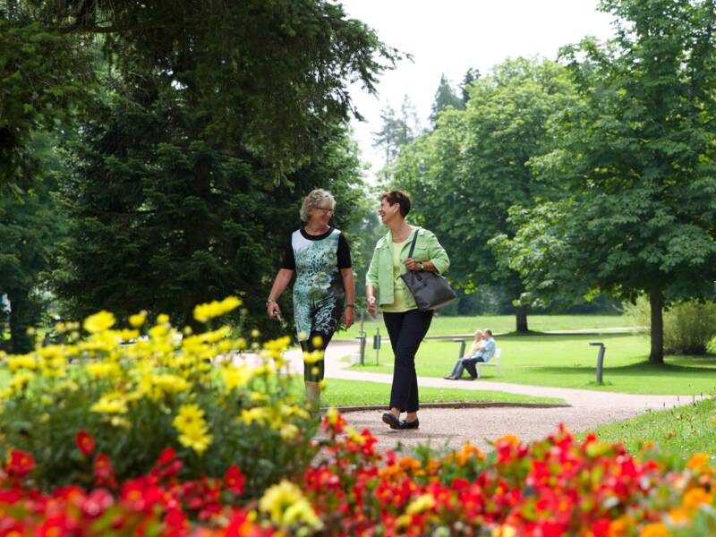 Zwei Frauen spazieren auf einem Weg durch einen blühenden Park mit vielen Bäumen und bunten Blumen.