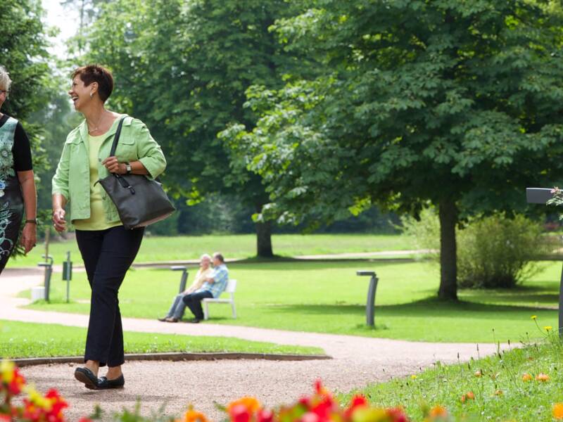 Zwei Frauen spazieren auf einem Weg durch einen blühenden Park mit vielen Bäumen und bunten Blumen.