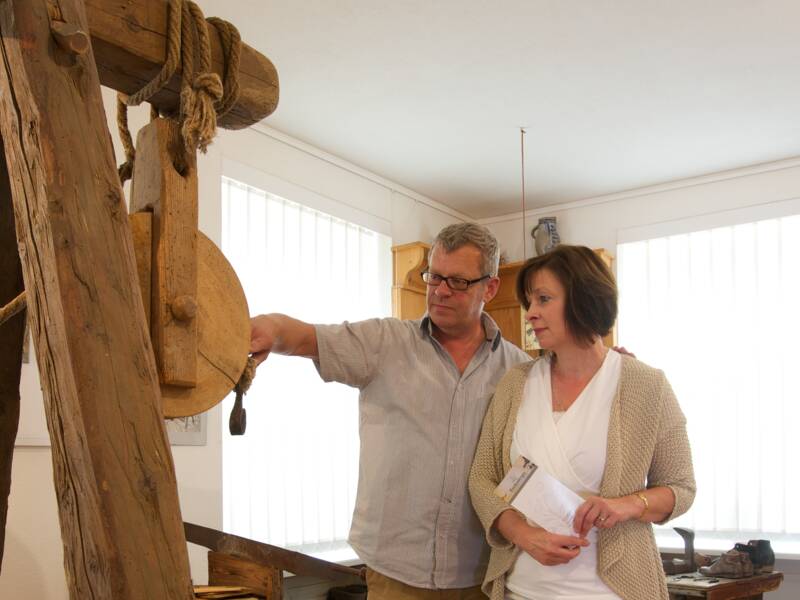 Zwei Personen betrachten in einem Museum eine historische Holzkonstruktion mit einem Flaschenzug, umgeben von alten Werkzeugen und Kleidungsstücken.