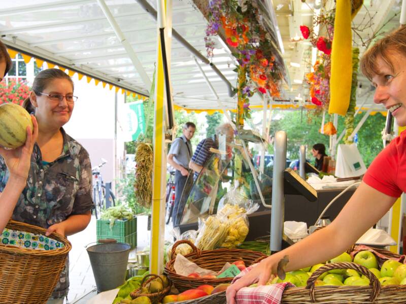 Drei Frauen stehen an einem Marktstand mit Obst und Gemüse. Eine der Frauen riecht an einer Melone. 