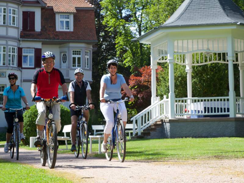 Zwei Männer und zwei Frauen fahren auf ihren Fahrrädern durch einen Park an einem weißen Pavillion vorbei. 