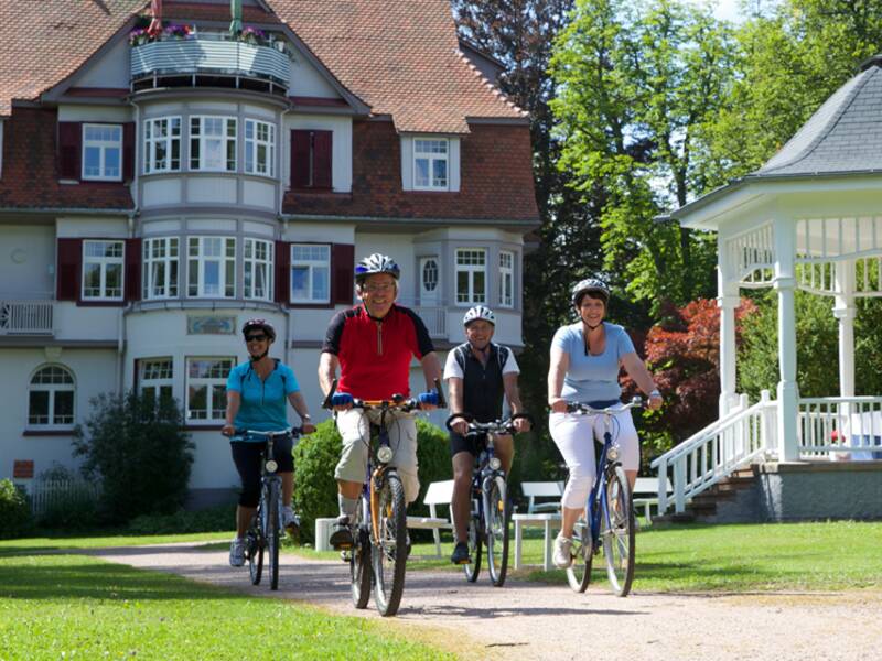 Zwei Männer und zwei Frauen fahren auf ihren Fahrrädern durch einen Park an einem weißen Pavillion vorbei. 