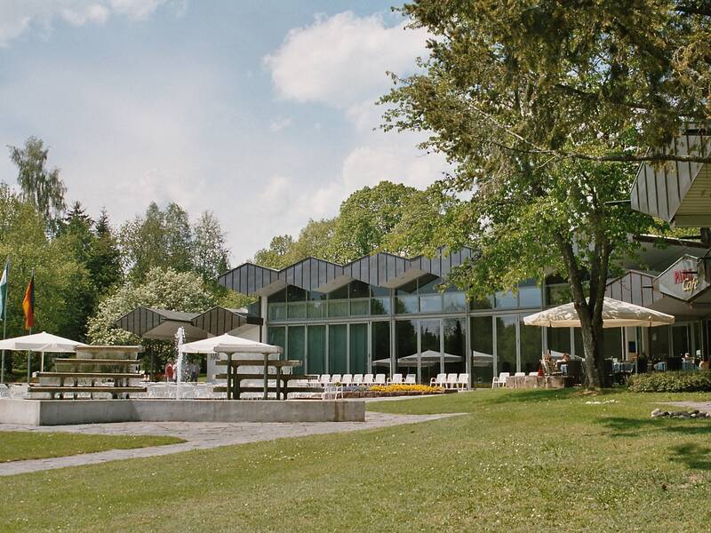 Ein modernes Gebäude mit großen Glasfenstern, umgeben von grünen Bäumen und Rasenflächen, mit mehreren Sonnenschirmen und Sitzgelegenheiten im Freien.