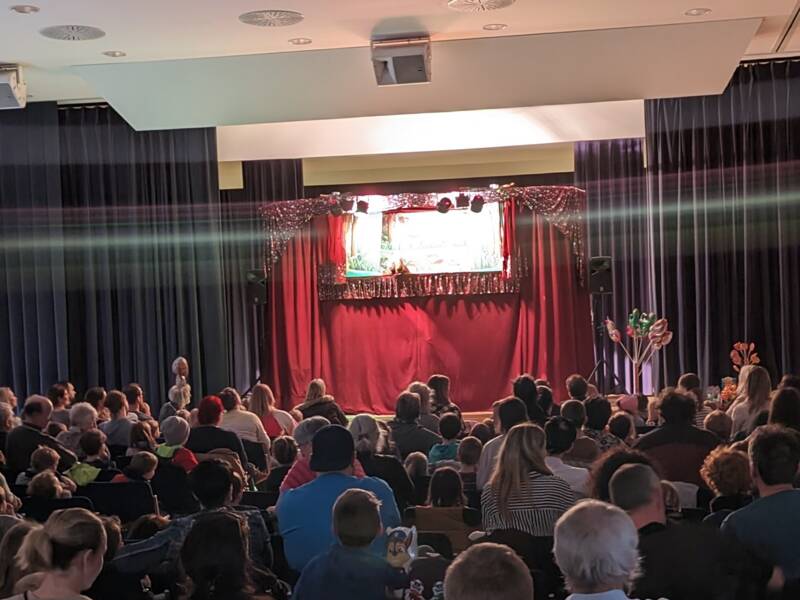 Eine große Menschenmenge sitzt in einem Saal und schaut auf eine Bühne mit einem roten Vorhang, auf der ein Puppentheater stattfindet.