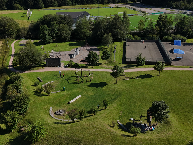 Luftaufnahme eines Parks mit Spielplatz, Grünflächen, Wegen, einem Fußballfeld im Hintergrund und Personen, die sich entspannen und spielen.