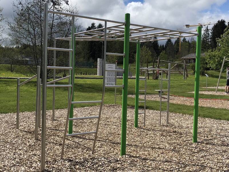 Ein Outdoor-Spielplatz mit Klettergerüsten und Fitnessgeräten ist von grünen Wiesen und Bäumen umgeben.