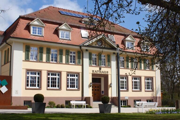 Ein zweistöckiges Rathaus mit beige-pinkfarbener Fassade, weißen Fensterläden und einem roten Ziegeldach, umgeben von grünem Rasen und einigen Bäumen unter klarem Himmel.