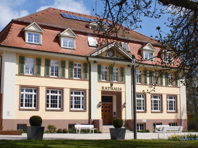 Ein zweistöckiges Rathaus mit beige-pinkfarbener Fassade, weißen Fensterläden und einem roten Ziegeldach, umgeben von grünem Rasen und einigen Bäumen unter klarem Himmel.