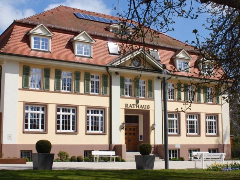 Ein zweistöckiges Rathaus mit beige-pinkfarbener Fassade, weißen Fensterläden und einem roten Ziegeldach, umgeben von grünem Rasen und einigen Bäumen unter klarem Himmel.