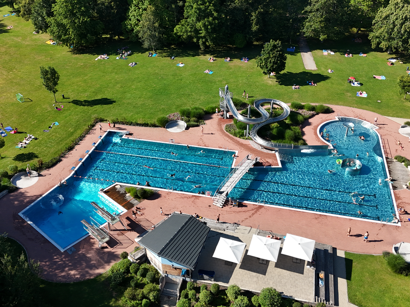 Das Bild zeigt eine Luftaufnahme eines belebten Freibads mit mehreren Schwimmbecken, einer Rutsche und sonnenbadenden Menschen auf der umliegenden Rasenfläche.