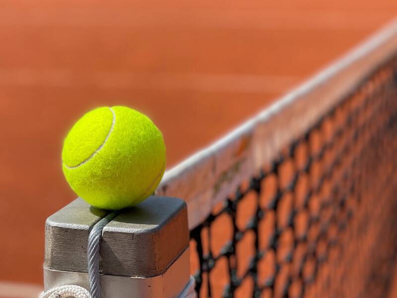 Ein Tennisball liegt oben auf dem Metallpfosten eines Tennisnetzes auf einem sandfarbenen Tennisplatz.