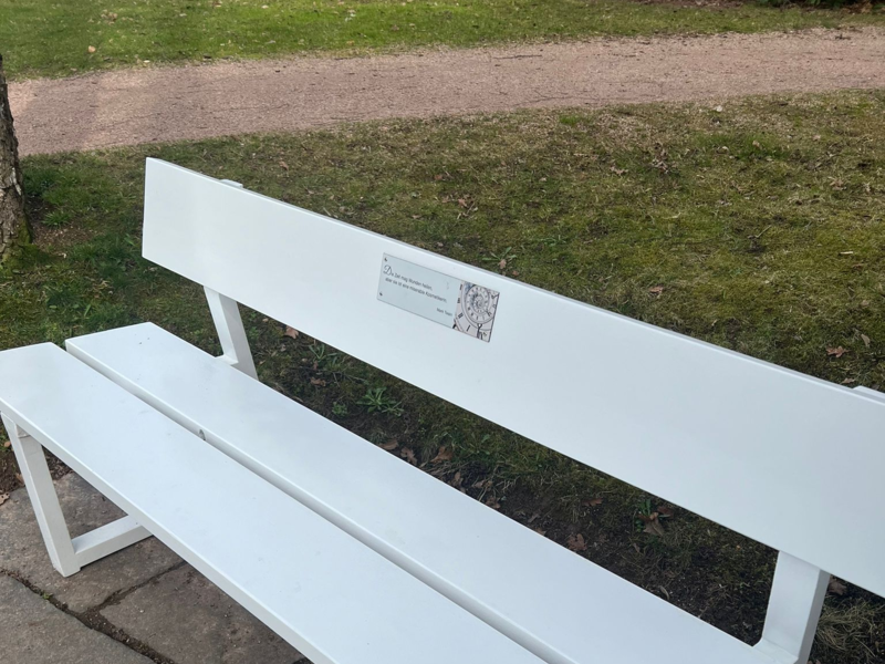 Weiße Parkbank aus Holz mit einer Rückenlehne, auf der ein Schild befestigt ist, steht auf einem gepflasterten Weg neben einer Grünfläche.