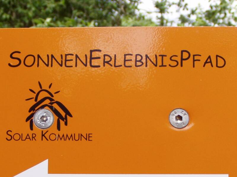 Ein orangenes Hinweisschild mit der Aufschrift "SonnenErlebnisPfad" und dem Zusatz "Solar Kommune" weist mit einem weißen Pfeil nach links.