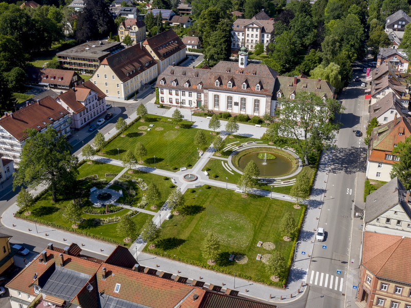 Luftaufnahme eines gepflegten Parks mit symmetrischen Wegen und einem zentralen Springbrunnen, umgeben von historischen Gebäuden und grünen Bäumen.