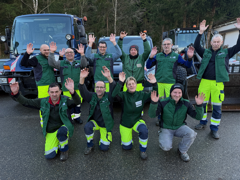 Eine Gruppe von jubelnden Männern und einer Frau in Arbeitskleidung in zwei Reihen vor drei Baufahrzeugen draußen auf einer Straße vor einem Tannenhintergrund.